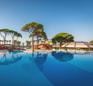 Gro&szlig;er, klarer Hotelpool mit modernen Pavillons und hohen Pinien im Hintergrund im Paloma Hotels Sencia Resort bei blauem Himmel.