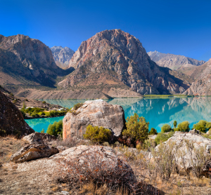 T&uuml;rkiser Bergsee spiegelt schroffe Gipfel der Fann-Berge, umgeben von Felsen und karger Vegetation.