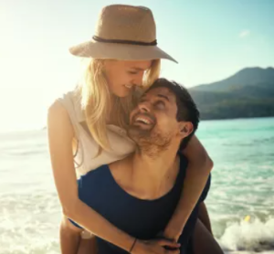 Lachendes Paar am Strand &ndash; sie tr&auml;gt einen Strohhut, er tr&auml;gt sie huckepack, im Hintergrund Meer und Berge.
