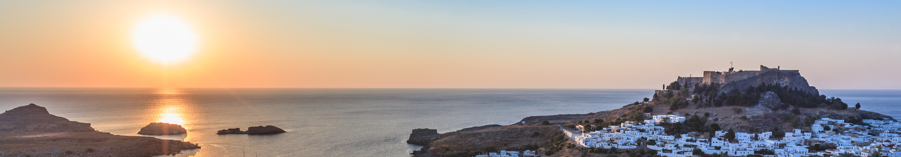 Sonnenuntergang &uuml;ber der K&uuml;stenstadt Lindos auf Rhodos mit Bucht, wei&szlig;en H&auml;usern und Akropolis auf einem H&uuml;gel.