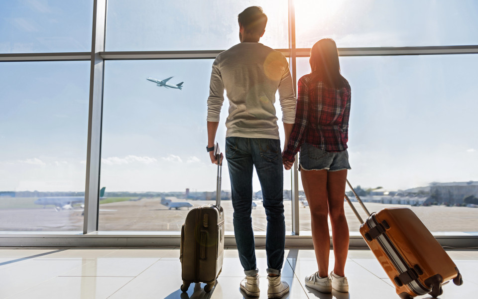 Reisepaar am Flughafen h&auml;lt H&auml;ndchen und blickt durch die gro&szlig;e Glasfassade auf ein startendes Flugzeug, bereit f&uuml;r ihr n&auml;chstes Abenteuer.