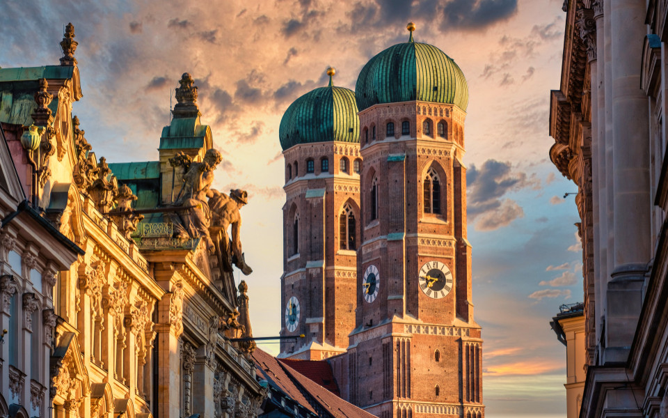 Sehensw&uuml;rdigkeit in der M&uuml;nchner Altstadt: Die M&uuml;nchner Frauenkirche mit ihren markanten Zwiebelt&uuml;rmen, eingerahmt von historischen Geb&auml;uden, leuchtet im warmen Licht der Abendd&auml;mmerung.