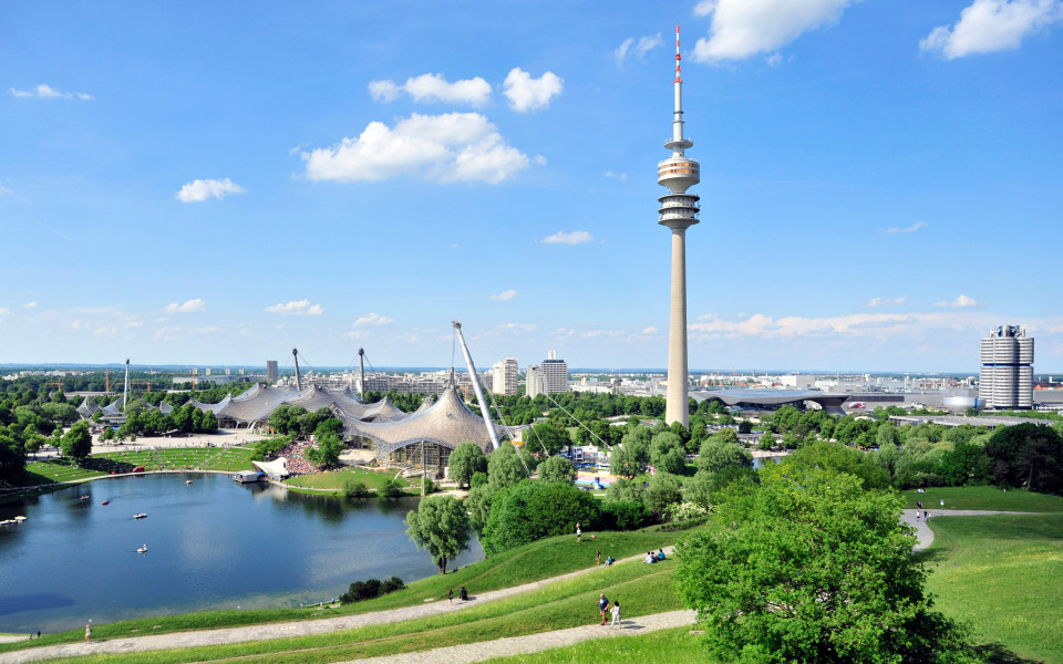 Geheimtipp in M&uuml;nchen: Der Olympiapark in M&uuml;nchen mit dem markanten Olympiaturm, der ikonischen Zeltdacharchitektur und dem idyllischen See, umgeben von gr&uuml;nen Wiesen unter blauem Himmel.