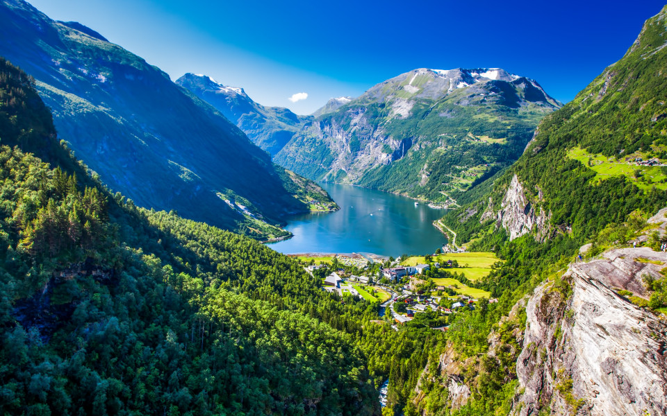 Der Geirangerfjord erstreckt sich tief zwischen steilen, gr&uuml;nen Bergh&auml;ngen mit schneebedeckten Gipfeln und einem kleinen Dorf am Ufer.